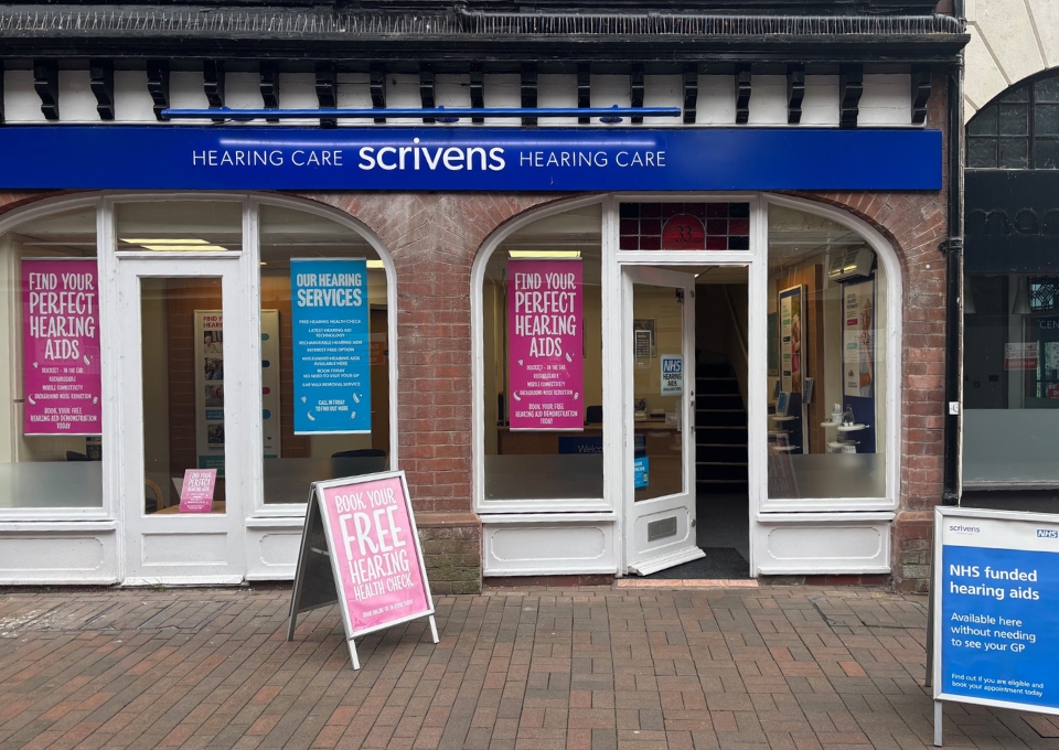 Shopfront of Scrivens Hearing Care with blue sign and arched windows showing posters about hearing aids and services outside the store
