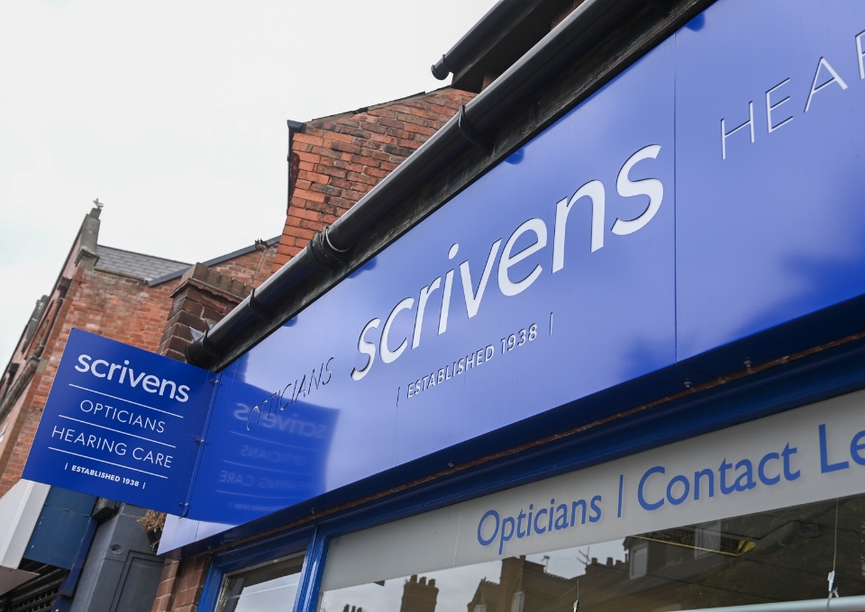 Blue storefront sign for Scrivens Opticians on a shopfront, with 'Opticians' and 'Contact L...' visible in the window