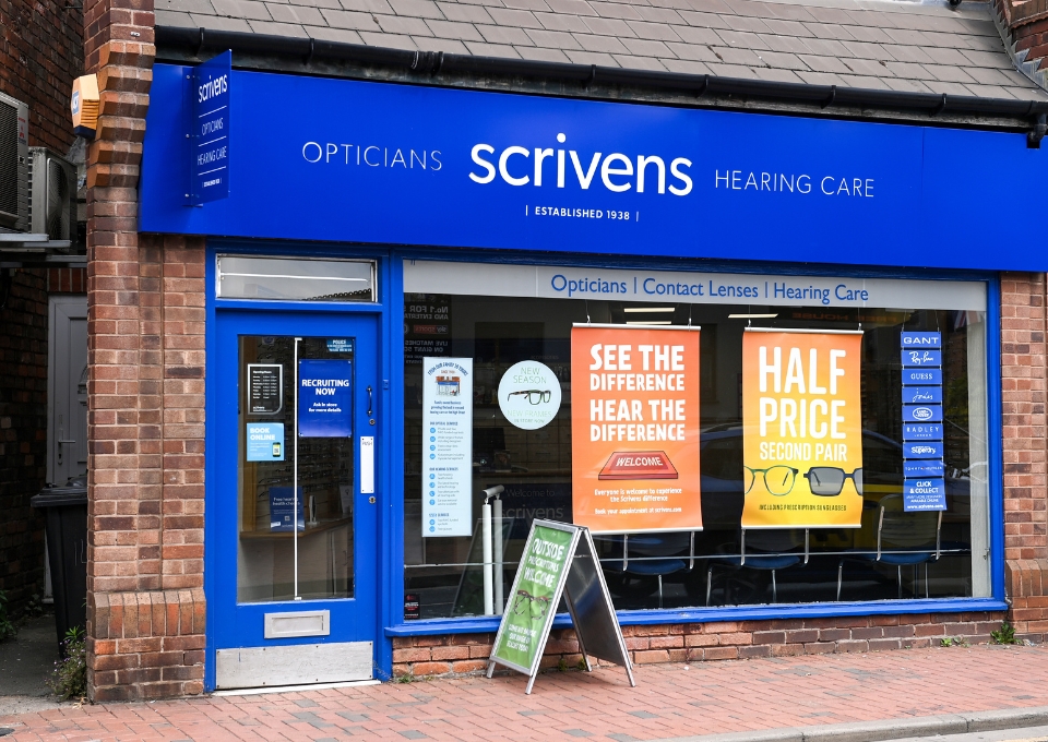 Blue storefront for Scrivens Opticians and Hearing Care with window posters promoting deals and services.