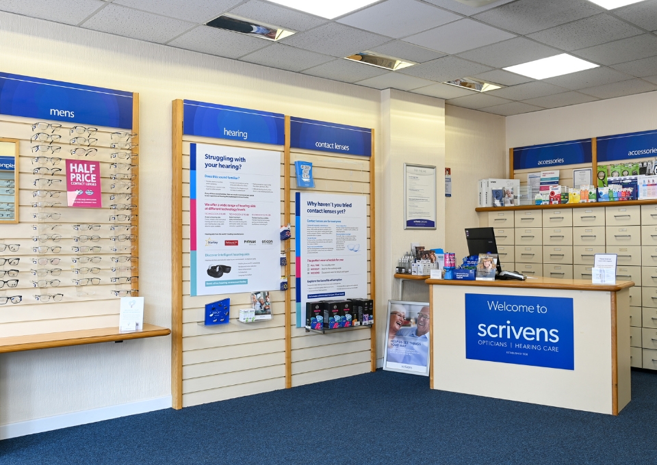 Bright optician shop interior with glasses on wall panels, posters about hearing and contact lenses, and a blue Scrivens reception desk.