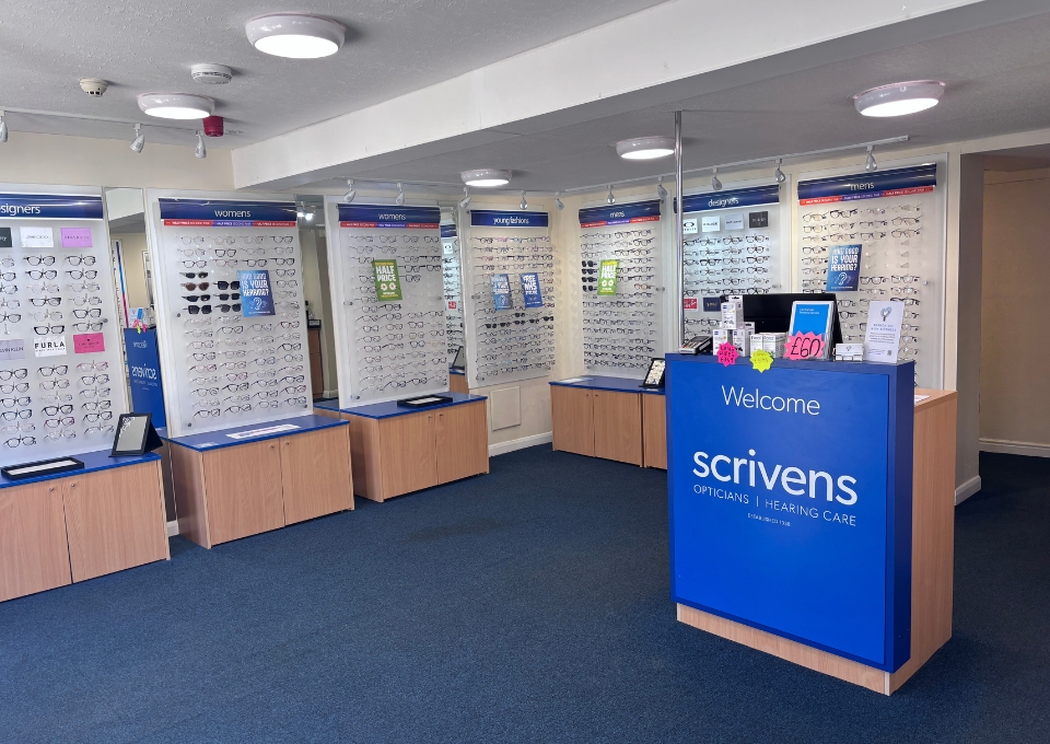 Interior of a Scrivens opticians shop with wall-mounted frame displays and freestanding display panels under track lighting, showcasing numerous eyeglasses.