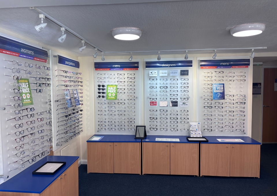 Eye doctor/optical shop interior with wall-mounted displays of eyeglasses for women and men, blue countertops, and wooden cabinets in the foreground.