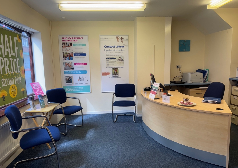 Reception area of a clinic with a curved light-wood desk, blue chairs, and posters about lenses on the wall—informational setting for visitors/appointments.