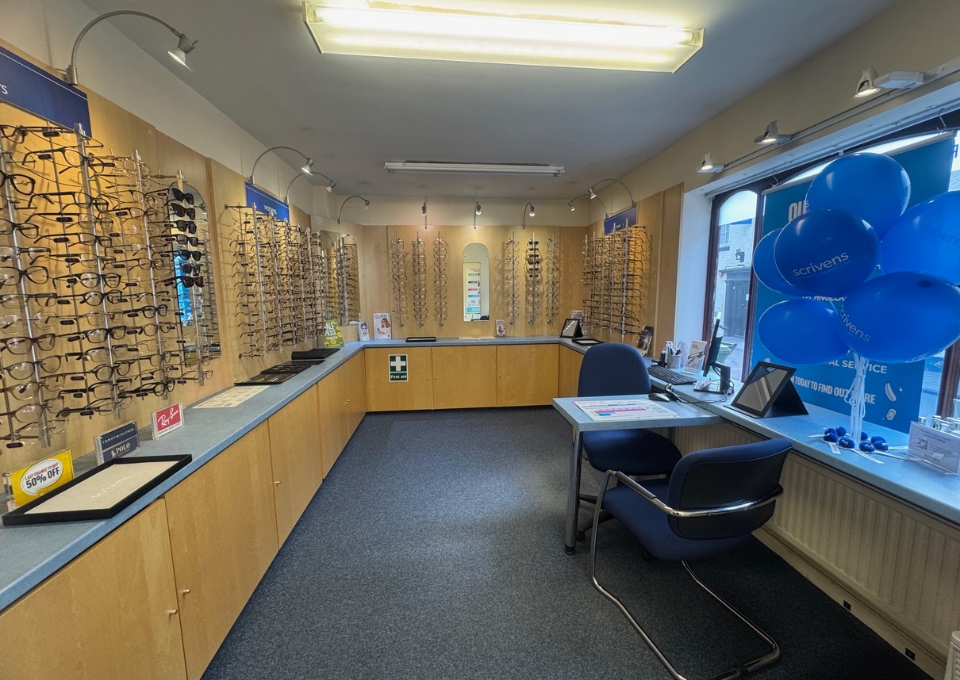 Optical store interior with wall-mounted eyeglasses displays, a long counter, and blue balloons near a service desk.