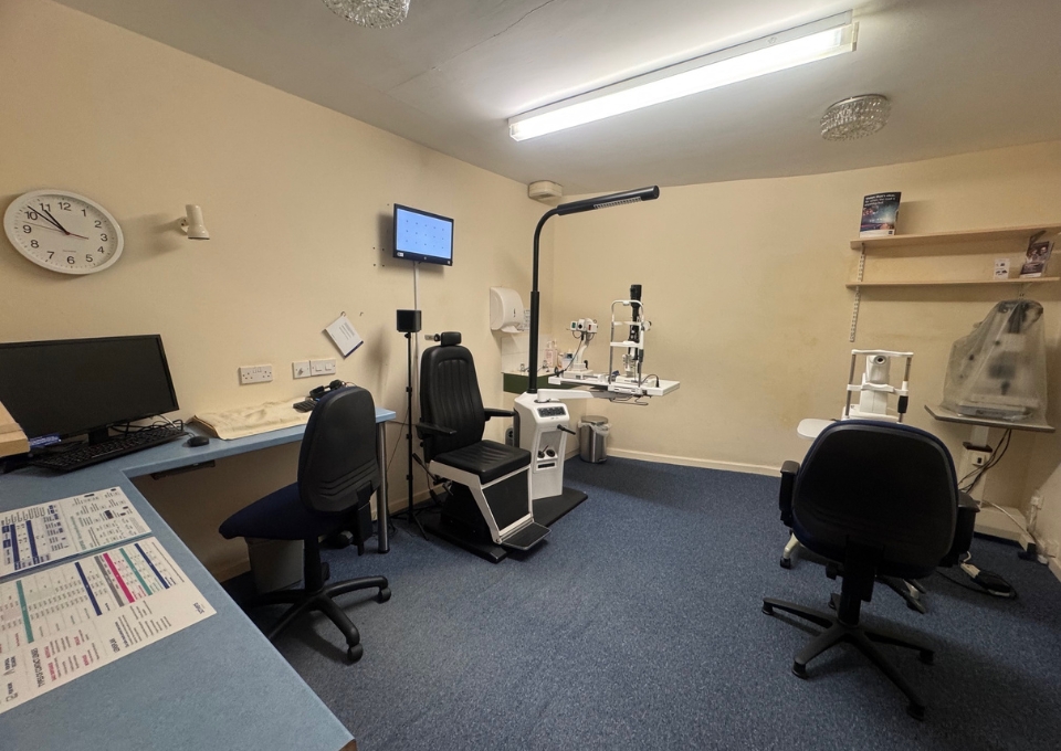Ophthalmology exam room with two chairs, a slit-lamp instrument, and diagnostic equipment on a blue-topped desk area.