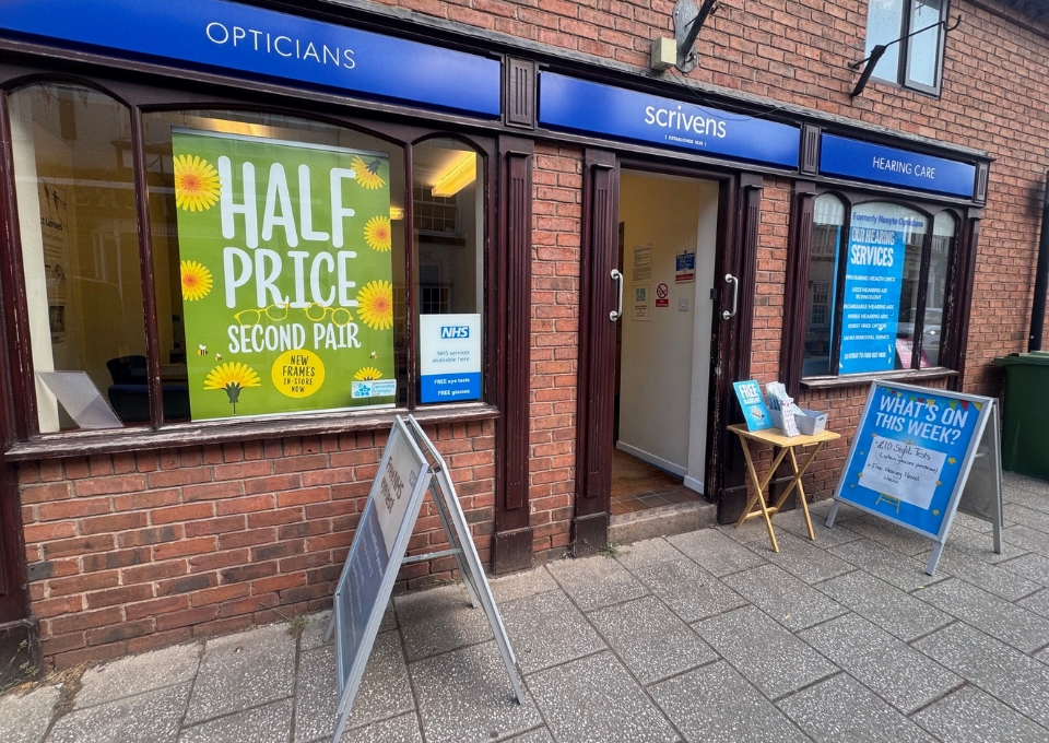 Shopfront with 'HALF PRICE SECOND PAIR' poster in the window; signs read Opticians and Scrivens Hearing Care on blue awnings and brick façade.