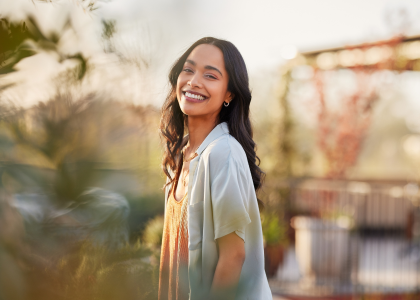 Young woman with long dark hair smiling in a sunlit outdoor setting in a garden or park atmosphere.