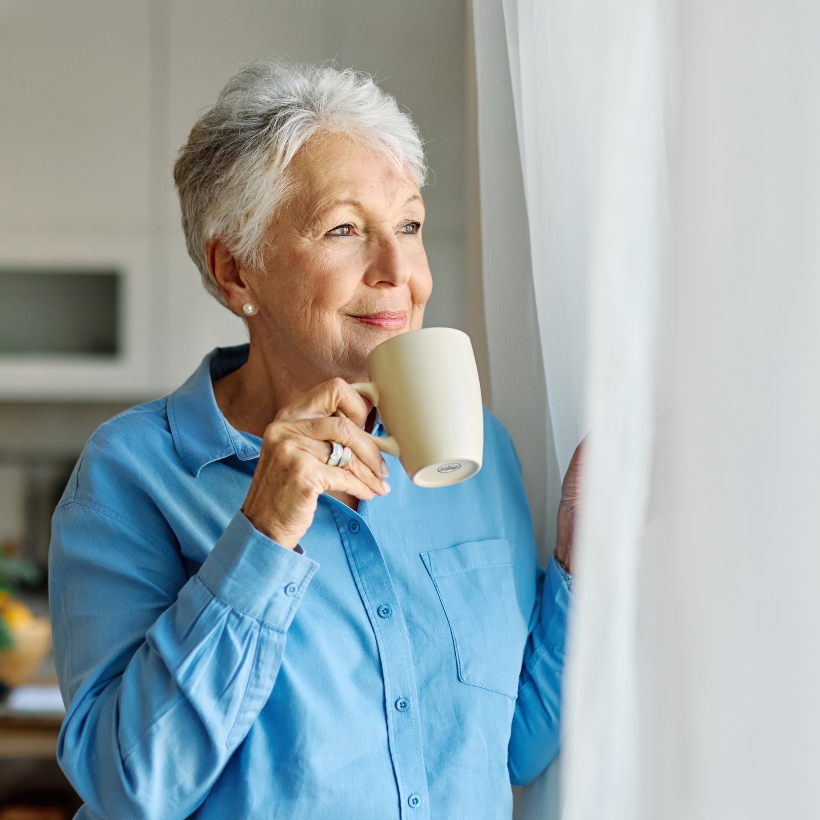 Older woman looking out curtains with a cup of tea