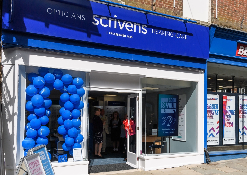 Blue Scrivens Hearing Care storefront with a large blue balloon arch outside and customers inside the open doorway, suggesting a special event or promotion.