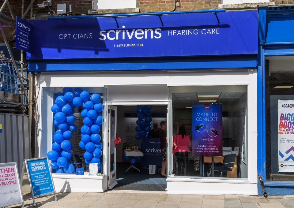 Blue Scrivens Hearing Care storefront decorated with blue balloons for a promotion/grand opening event nearby.
