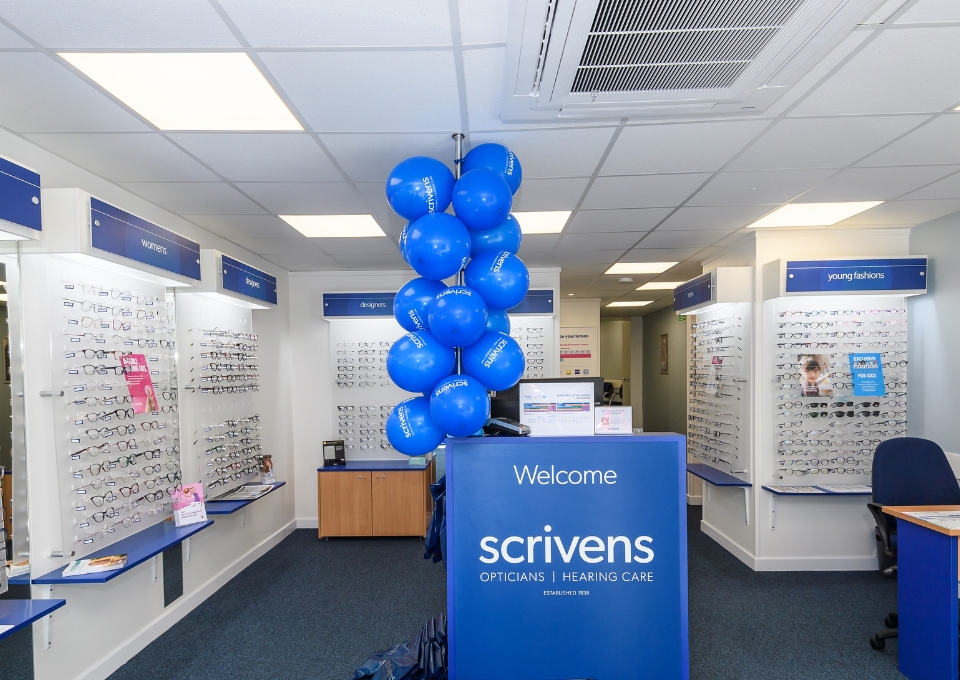 Reception desk with blue Scrivens sign and a column of blue balloons in an optician’s showroom, with eyeglasses displayed on walls to the left and right.