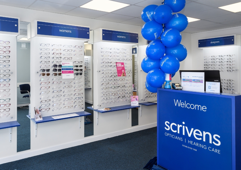 Eyewear store interior with wall displays of glasses labeled women's and designers; blue balloons cluster near a welcome desk that reads Scrivens Opticians.