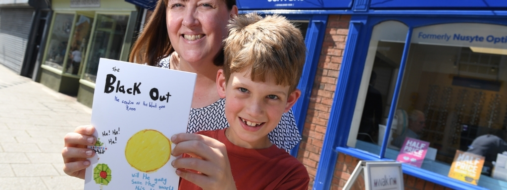 Woman and boy smile while holding a book titled 'The Black Out' in a sunny street with blue shop fronts.